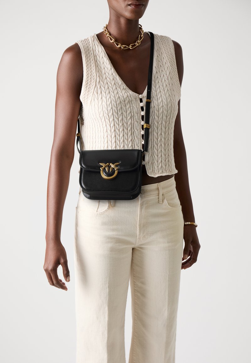 Black leather crossbody bag with a gold hardware detail, featuring a circular clasp and a textured finish, worn over a cream knit top.