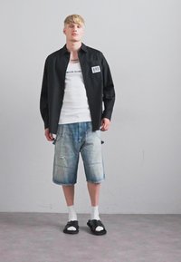 Blond man med svart knappad skjorta, vit linne, slitna denimshorts, vita strumpor och svarta sandaler, som står mot en enfärgad grå vägg.