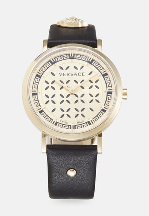 Reloj tono dorado con correa de cuero negro, dial con patrón y recortes, diseño de clave griega alrededor del borde, etiqueta de fabricación suiza.