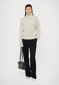 Pull à col roulé en maille tissée beige clair, pantalons noirs à rayures, et un sac à main bleu marine avec une boucle. Le modèle se tient devant un fond neutre.