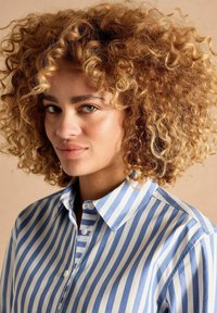 Striped button-up shirt in light blue and white, featuring a collar, front placket, and textured fabric. Hair is voluminous and curly.