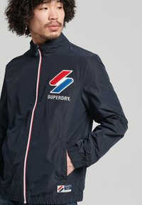Chaqueta cortavientos de color marino con un alto cuello, con logotipos bordados en rojo y azul, cierre de cremallera y puños elásticos; dos bolsillos laterales.