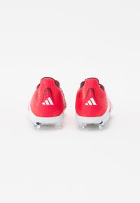 Chaussures de soccer rouges avec un matériau texturé, des accents blancs et un logo à trois bandes à l'arrière, offrant un design léger et des crampons transparents.