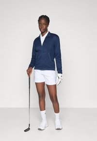 Young woman in navy jacket, white golf gloves, shorts, and sneakers holding a golf club, standing against a plain white background.