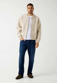 Beige bomber jacket with ribbed collar and cuffs, worn over a white t-shirt. Paired with dark blue straight-leg jeans and brown slip-on shoes.
