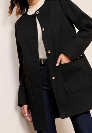 Mujer con un abrigo negro con botones dorados, top beige, vaqueros oscuros y cinturón negro, con una mano en el bolsillo del abrigo.