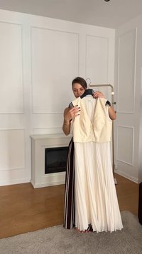 Elfenbeinfarbenes ärmelloses Vest mit zwei Fronttaschen und Knöpfen, kombiniert mit einem plissierten cremefarbenen Rock, gehalten von einer Person in einem gestreiften Kleid.