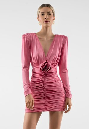 Žena ve světle růžových saténových nařasených minišatech s dlouhými nabíranými rukávy, hlubokým výstřihem do V a velkým růžovým detailem v pase.