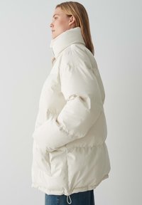 Giacca a vento, bianca, con colletto alto e tasche laterali. Presenta una texture trapuntata e un orlo elastico. Abbinata a jeans blu, vista di profilo laterale.