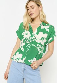 Camicia button-up verde con motivo floreale bianco, maniche corte e vestibilità rilassata, abbinata a jeans di denim blu chiaro.