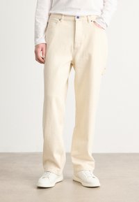 Pantalons en coton beige avec une coupe droite, dotés de boutons bleus, de poches latérales et d'une texture lisse. Associés à des baskets blanches.