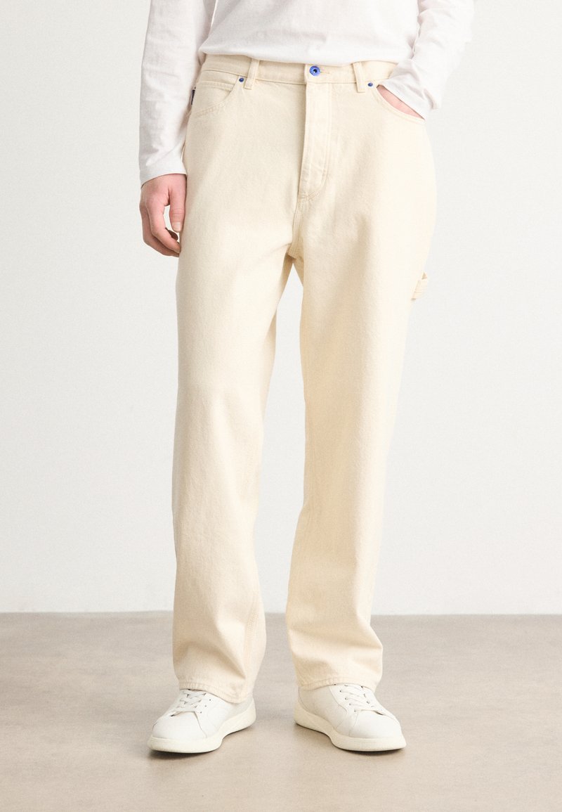 Pantalons en coton beige avec une coupe droite, dotés de boutons bleus, de poches latérales et d'une texture lisse. Associés à des baskets blanches.
