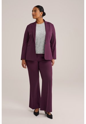 Blazer su misura violaceo e pantaloni a gamba larga coordinati con un maglione in maglia grigia chiara sotto; scarpe con tacco a punta nere.