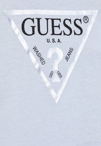 Pilka medvilninė marškinėlių su dideliu juodai baltu trikampiu Guess logotipu, į kurį integruoti žodžiai "WASHED" ir "JEANS".