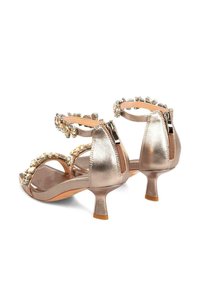 Tacones abiertos de piel metálica dorada con cremallera en la parte posterior, correas adornadas y un tacón modesto. Textura suave con detalles florales en la correa.