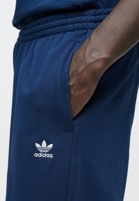 Tmavomodré tepláky z bavlneného zmesi s elastickým pásom. Majú bočnú kapsu a bielu logo Adidas na dolnej časti nohy.