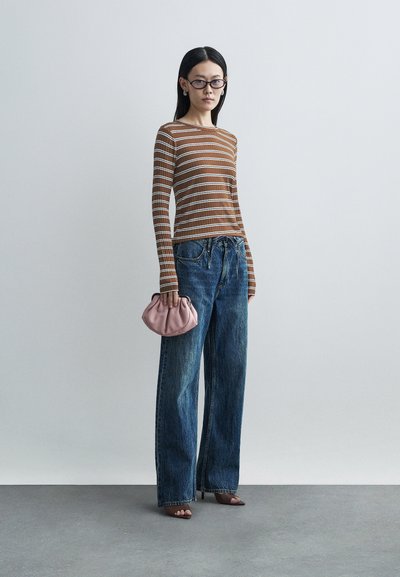 Top de manga comprida com riscas castanhas, jeans azuis largos e uma mala de mão rosa. Modelo a usar sandálias castanhas com tiras, em pé sobre um chão cinzento.