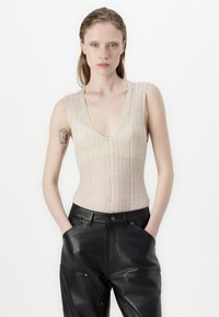 Teksturowany, jasnobeżowy dzianinowy top na ramiączkach z głębokim dekoltem w serek, noszony z czarnymi skórzanymi spodniami z wyraźnymi kieszeniami i złotymi detalami.