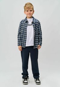 Niño vestido con una camisa de cuadros azul y blanca sobre una camiseta blanca con un gráfico, combinada con pantalones deportivos marinos y zapatillas negras.