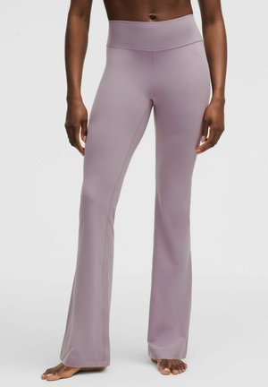 Vrouw draagt hoge, mauvekleurige uitlopende yogabroek, staat blootsvoets tegen een effen lichte achtergrond.