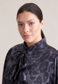 Schwarze Satinbluse mit einer Schleifenkragen und Leopardenmuster. Der Stoff hat eine glatte Textur und einen dezenten Glanz.