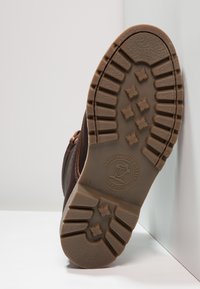 Sapata de couro castanho com uma sola de borracha texturizada, apresentando um padrão de ranhura e detalhe de logo. A sapatilha é exibida a partir da vista da sola.