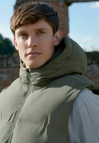 Olivengrüne gepolsterte Weste mit hohem Kragen und Reißverschluss, die eine glatte Textur und strukturierte Schultern aufweist, getragen über einem hellgrauen Pullover.