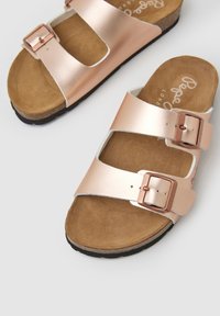 Roze gouden sandalen met twee brede metalen banden en verstelbare gespen, voorzien van een zachte suède-achtige inlegzool en een gestructureerde zwarte buitenzool.
