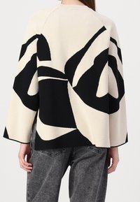 Femme portant un pull crème avec de grandes formes abstraites noires audacieuses, associées à un jean noir délavé, vue de dos sur un fond blanc.