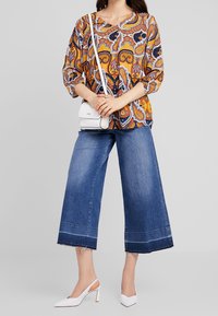 Kvinde iført farverig paisley-bluse, brede ankelbukser, hvide slingback hæle, og holder en lille hvid crossbody taske.