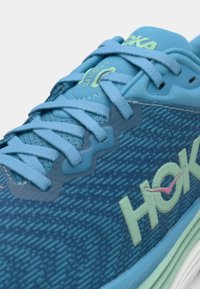 Chaussure de course bleue avec une tige en mesh, motif texturé, lacets et marque HOKA. Accentuée de détails vert clair et rose.
