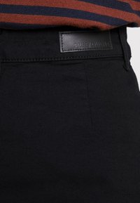 Černé kalhoty s hladkou texturou, opatřené černou koženou cedulkou s ražebním textem. Tkanina je mírně elastická, což zvyšuje pohodlí.