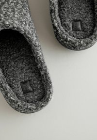 Graue Wollslipper mit strukturiertem Oberflächenmaterial, runder Form und flacher Sohle. Enthält ein kleines, gebrandetes Lederetikett im Inneren.