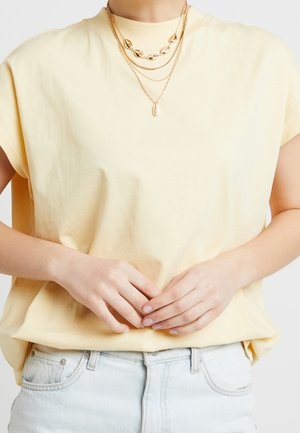 Femme portant un haut sans manches jaune pâle, des colliers en or superposés et un jean clair, les mains délicatement jointes à la taille.