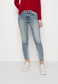 Jeans ajustados de un azul claro con un acabado desgastado, diseño de cinco bolsillos y corte a la altura del tobillo, combinados con un suéter texturizado rojo y zapatillas grises.