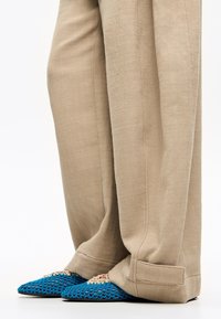 Blauwe geweven mesh schoenen met spitse neuzen, voorzien van beige veters, gecombineerd met beige linnen broeken. De schoenen liggen op een gladde ondergrond.