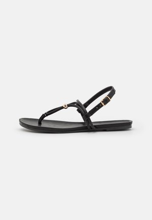 Sort flad sandal med tynde remme, en guldbelik ved anklen og en guldkugledetalje på remmen ved tåen.