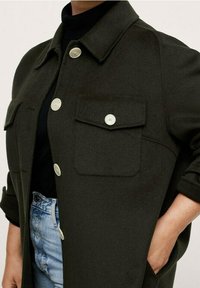 Chaqueta de mezcla de lana verde oscuro con cuello de camisa, dos bolsillos frontales y botones mate. Estilizada sobre una blusa negra y jeans de un azul claro.