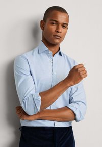 Camicia a quadri blu chiaro con colletto a punta, maniche arrotolate e silhouette aderente. Il tessuto appare liscio e strutturato.