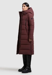 Abrigo largo de plumas burdeos con capucha, con acolchado horizontal, bolsillos con cremallera y silueta ajustada, combinado con botas negras hasta la rodilla.