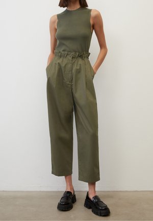 Mujer con top verde oliva sin mangas, pantalones verdes oliva sueltos de tiro alto con bolsillos y mocasines negros gruesos, de pie en el interior.