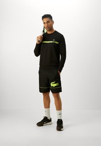 Sweatshirt preto com logótipo da Lacoste em várias cores, combinado com calções pretos que apresentam um emblema de crocodilo verde e detalhes fluorescentes.