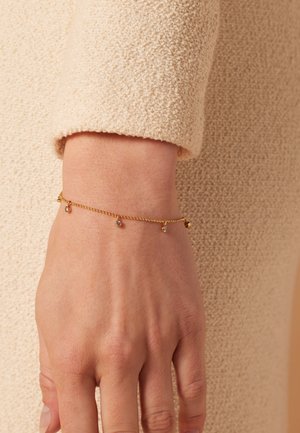 Gouden kettingarmband met kleine edelstenen en delicate belaccenten, weergegeven op een blote pols tegen een gestructureerde beige achtergrond.