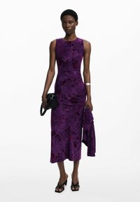 Robe à imprimé floral violet avec une silhouette ajustée, un col rond et une longueur jusqu'aux genoux. Le modèle tient un sac à main noir et porte des sandales noires.
