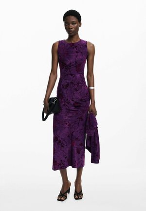 Robe à imprimé floral violet avec une silhouette ajustée, un col rond et une longueur jusqu'aux genoux. Le modèle tient un sac à main noir et porte des sandales noires.