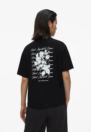 Osoba o ciemnych włosach, ubrana w czarną koszulkę z białym napisem „Karl Lagerfeld Jeans” i abstrakcyjną grafiką na plecach.
