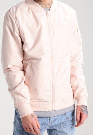 Personne portant une veste bomber rose clair avec une fermeture éclair à l’avant et un pantalon bleu avec des motifs abstraits noirs, les mains détendues le long du corps.