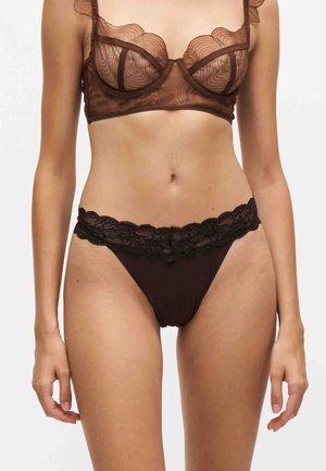 Femme portant un soutien-gorge en dentelle fine marron et une culotte assortie avec une taille en dentelle festonnée, debout devant un fond uni.