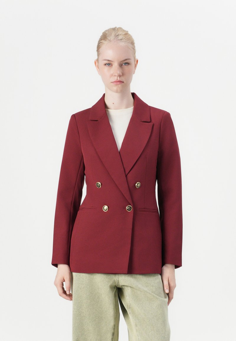 Bordeaux dobbeltradet blazer med gullfargede knapper, strukturerte skuldre og slank design. Båret over en kremfarget topp og lysegrønne bukser.