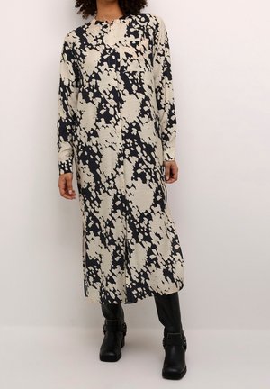 Femme portant une robe midi à manches longues, boutonnée, avec un motif abstrait noir et beige, ainsi que des bottines noires, debout devant un fond uni.